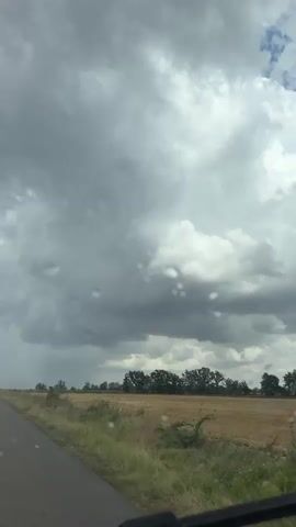 This is a short video shot from inside a vehicle, looking out through the windshield at a rural landscape. The sky is filled with dramatic, dark, and heavy clouds, suggesting an impending storm. There are raindrops visible on the windshield, and some streaks from the windshield wipers can be seen. The road is visible in the lower left portion of the frame. Beyond the road, there is a field of dry, golden stubble, likely harvested crops. A line of trees stands against the horizon. The overall mood is somber and atmospheric, emphasizing the approaching weather.