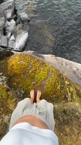 A first-person perspective video shows a person's bare feet standing on moss-covered rocks next to a body of water. The person is wearing a light-colored skirt. The water is calm with ripples on the surface. The rocks are gray and rough, with patches of green moss. The scene appears to be a natural outdoor environment.
