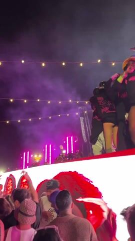 The video shows a live music performance with a crowd of people watching and recording with their phones. The stage is brightly lit with pink and white lights and has a large LED screen displaying faces. Performers are on stage, some with unique headwear, dancing and interacting with each other. String lights are hung overhead, and there is smoke or haze in the air. People in the crowd are dressed casually, with some wearing hats.