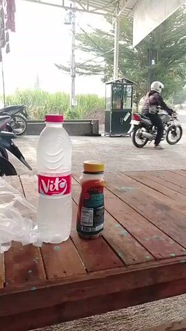 A static, eye-level shot focuses on a weathered wooden table with two plastic bottles and a white plastic bag on it. The larger bottle, clear and filled with water, has a red cap and a red and white label that reads "Vito." Next to it, a smaller bottle, amber-colored and likely containing a beverage, has a brown cap and a label with visible text. The plastic bag is partially opened. In the background, out of focus, a person wearing a white helmet rides a motorcycle from left to right. Beyond the motorcycle, a street and a small building with glass panes can be seen. The environment appears to be outdoors, possibly a roadside stall or rest area, with overhead structures and vegetation visible in the distance. The lighting suggests daylight, though the sky is overcast.