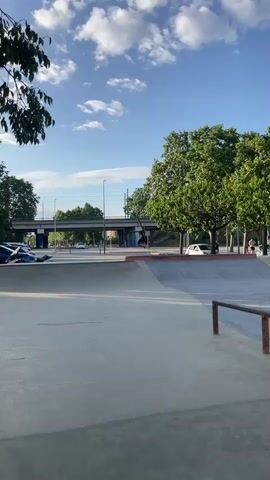 The video shows an outdoor skatepark during what appears to be late afternoon or early evening, indicated by the long shadows and bright sunlight. The sky is clear with a few scattered clouds. The skatepark features concrete ramps, bowls, and metal rails. Green trees line the perimeter of the park, and a road with cars is visible in the background, along with an elevated bridge or overpass. Several people are present, some walking with skateboards, others sitting or standing around the park. One person walks towards the camera holding a skateboard. Graffiti is visible on some of the ramps, including the word "LIBIADS" and a cartoon character with "ZALO" nearby. The overall atmosphere seems relaxed and active, typical of a community skatepark.