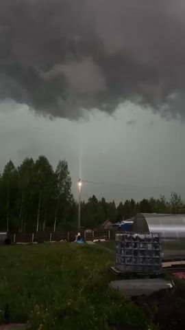 A wide shot shows a yard with a grassy field, a dark wooden fence, trees, a small shed, a greenhouse, and several other structures under a sky heavy with dark gray clouds. A single streetlight is on, casting a bright beam into the ominous atmosphere. A blocky metal container sits in the foreground. There is slight movement in the leaves of the trees, suggesting a breeze. The overall tone is somber and suggestive of an impending storm.