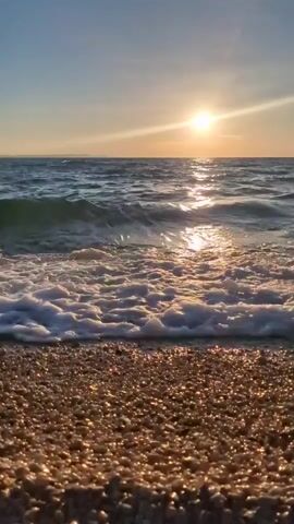 The media depicts a serene beach scene at sunset in Zadar, Croatia. The setting is a pebble beach with the calm, dark blue sea stretching towards the horizon. The sun, a bright orb with visible rays, is setting in the sky, casting a warm, golden glow on the water and the wet pebbles. Gentle waves are rolling onto the shore, creating white foam and reflecting the sunlight. The sky is a clear, pale blue, transitioning to a warmer hue near the horizon where the sun is positioned. The overall mood is peaceful and tranquil, evoking a sense of calm and natural beauty. There is no visible text in the media.