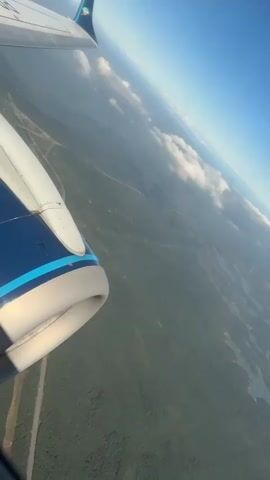 A vertical-format aerial shot from a plane window looks down on a landscape of water and wooded land. The plane wing and engine are visible in the upper-left of the frame. The plane appears to be banking slightly, as the horizon is tilted. The sky is bright and blue with some scattered clouds.