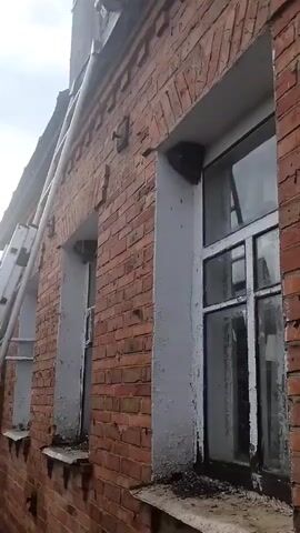 A camera is positioned looking upwards at the side of a brick building. Two windows are visible. To the left of the right-hand window, in the upper corner, a dark, round object appears to be attached to the brickwork. A ladder leans against the building to the left. A cloudy sky is visible above the building.