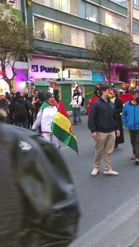 The video depicts a street scene with a group of people and motorcyclists. The people are carrying and wearing the flag of Bolivia. The motorcyclists are lined up in the street, some wearing helmets and jackets.  The buildings lining the street have signs for businesses such as a pharmacy, a bank, and a telecommunications company. There are trees on the sidewalk. The light indicates it is daytime, likely early evening. The mood appears to be patriotic, with people displaying national symbols.