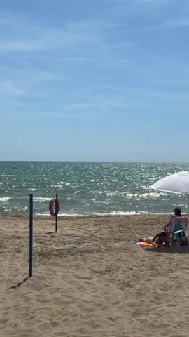 A sunny beach scene shows the ocean with sparkling waves under a clear blue sky. In the foreground, sandy beach stretches across the frame. On the left, a blue pole stands in the sand with a red and white lifebuoy attached. On the right, a white beach umbrella provides shade for two individuals. One person is seated in a multicolored beach chair facing the ocean, while another person lies prone on a colorful towel on the sand. The camera slowly zooms in on the ocean.