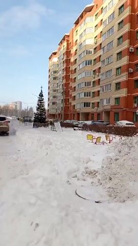 The video shows a snowy courtyard in a residential area on a bright, sunny day.  Tall apartment buildings border the courtyard on both sides. In the center, there's a small, decorated Christmas tree near a playground. A gold-colored SUV drives slowly along the snowy path. Later, two people, possibly a child and an adult, walk together in the snow.  Piles of snow are visible, and other cars are parked along the sides.