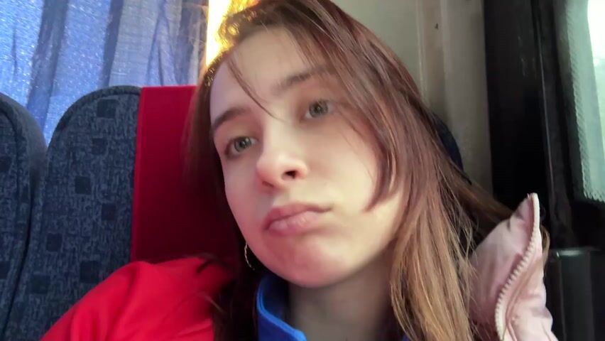 A young woman with light skin and light brown hair sits on a bus. She wears a red jacket over a blue top and a light pink padded jacket. She opens her mouth as if yawning, then holds up a peace sign. The seat next to her is covered in a blue and gray patterned fabric with a red headrest. A curtain hangs in the window behind her.