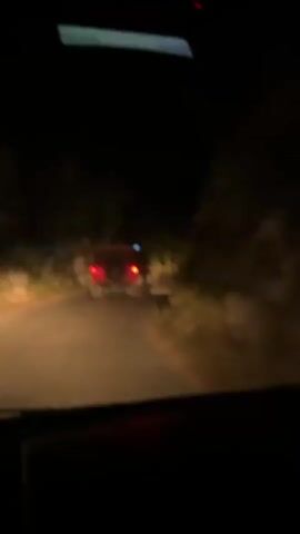 A red Jeep Wrangler is driving on a paved road at night. The Jeep is in front of the camera's vehicle, and its taillights are visible. The road is lined with trees and vegetation. The camera vehicle's headlights illuminate the road and the surrounding area. The Jeep has a spare tire mounted on the rear. The Jeep has blue and red lights on top.