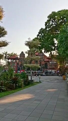 The video shows a wide paved walkway leading towards a large, ornate building with traditional architecture, likely a temple or palace, characterized by tiered roofs and intricate carvings. Lush green trees and foliage frame the scene, casting shadows on the walkway. In the background, a crowd of people and parked motorcycles are visible, suggesting a public space or event. The sky is clear and bright, indicating daytime. The camera is stationary and captures a long shot of the environment.