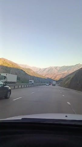 This is a first-person perspective video taken from inside a car driving on a multi-lane highway. The highway is surrounded by mountainous terrain. The sky is clear and appears to be late afternoon or early morning due to the lighting on the mountains. Several other vehicles, including cars and trucks, are visible on the highway, some in front and some in the opposite direction. The car is traveling forward on the right lane of the highway. The video appears to be a continuous shot with no cuts or edits.