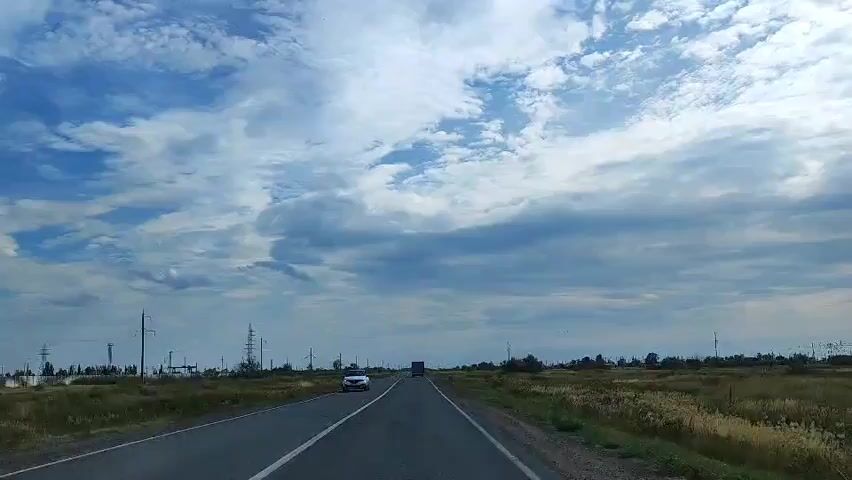 This is a first-person view from a vehicle driving down a paved road. The sky is mostly blue with scattered white and gray clouds. On either side of the road, there are grassy fields with some dry vegetation and scattered trees or bushes. Utility poles with power lines are visible along the left side of the road. In the distance, there are a few buildings and more utility poles. A white car is visible on the left side of the road, partially in frame. A truck is visible in the distance, driving in the same direction as the camera vehicle. The road has white dashed lines marking the lanes.