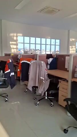 The video shows an empty office space with cubicles. The cubicles have desks with chairs. Some chairs are covered with blankets. There are windows that let in sunlight. There is a water cooler in the corner. There are papers and office supplies on the desks. The room is well-lit and appears to be clean. The video pans around the room, showing different angles of the cubicles and desks.