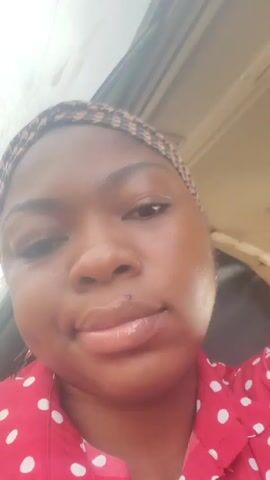 A woman is in a vehicle and looking at the camera. She is wearing a red and white polka dot shirt and a patterned headscarf. She makes a kissing face at the camera. The video is taken at an angle from above.