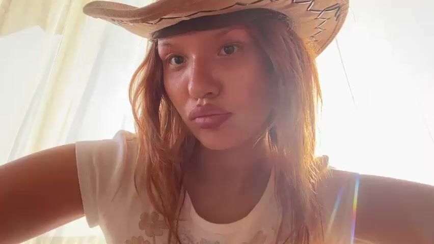 A young woman with red hair and a cowboy hat on is looking into the camera. She is wearing a white t-shirt. The background is a curtain. The lighting is bright and appears to be from a window. She is making different facial expressions and gestures with her hands.