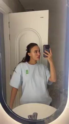 A woman is taking a selfie in a bathroom mirror. She has light brown skin, dark hair pulled back with a hair clip, and light-colored eyes. She is wearing a light blue t-shirt with a small green logo on the left side of her chest and a silver necklace. Her face has a clear, wet substance on it, possibly a face mask or moisturizer. She is holding a black phone in her right hand, presumably to take the picture. The background includes a white door with an arched panel, a white sink, a silver faucet, and patterned tile on the wall. The mirror has an oval shape with a dark blue frame and integrated lighting.