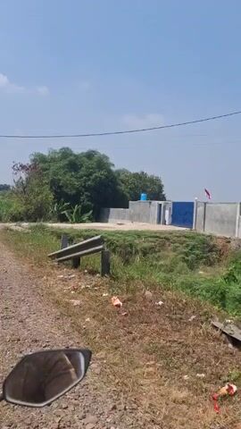 A first-person perspective video captures a view from a moving vehicle, likely a motorcycle, on a sunny day. The road is paved on the left, with a narrow dirt shoulder on the right. A metal guardrail runs along the edge of the shoulder, separating it from a ditch filled with green grass and some debris. In the distance, a building complex is visible, featuring concrete walls, a blue door, and a bright blue water tank on the roof. Lush green trees and vegetation surround the area. The sky is clear and blue with a few wispy clouds. The overall environment appears to be rural or semi-rural.