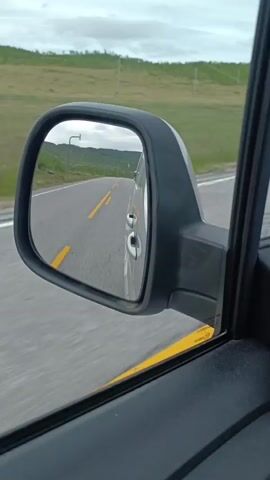 The video shows a view from inside a vehicle, focusing on the side mirror. The mirror reflects a highway with yellow double lines stretching into the distance. The landscape outside the car appears to be rolling hills covered in green vegetation, under a cloudy sky. The perspective suggests the vehicle is in motion, traveling along the road. No people are visible, and the primary objects are the car's interior and the reflection in the side mirror.