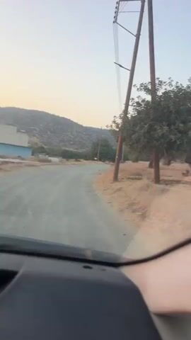 The video shows a view from inside a moving vehicle, looking out a side window and through a side mirror. The landscape outside is dry and grassy, with scattered trees. The sky appears clear and pale. The vehicle is on an unpaved road.