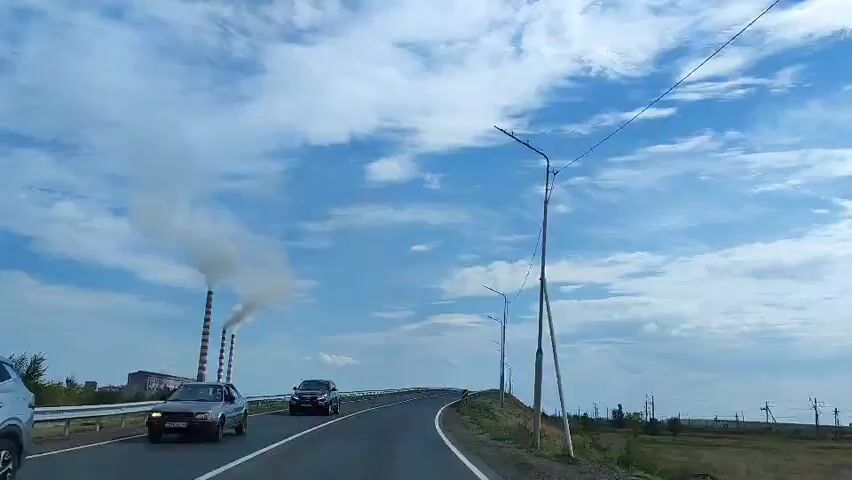 A black SUV drives on a highway. The highway is paved and has white lane markings. There is a guardrail on the right side of the road. The sky is blue with white clouds. In the distance, there are industrial smokestacks emitting smoke. There are streetlights on the right side of the road.
