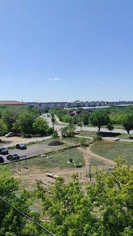 This is a vertically oriented, wide-angle video recording taken from a high vantage point, likely a balcony or window of a multi-story building. The scene depicts a residential area with a clear blue sky and a bright, sunny day, suggesting it's midday.

The foreground on the right is dominated by lush green foliage, indicating the presence of trees, possibly in a park or residential courtyard. To the left, a cityscape unfolds. A road runs through the scene, lined with apartment buildings of varying heights and architectural styles. Many of these buildings appear to be multi-story, typical of Soviet-era construction, with rows of windows visible. The road is partially lined with parked cars, suggesting residential activity. A dirt path also winds through the grassy areas, hinting at recreational spaces or informal walking routes.

In the middle ground, there are visible signs of a children's play area, with what appear to be old tires used for some kind of activity or decoration. There are also some wooden structures that could be part of playground equipment. The ground is mostly dry and dusty in these areas, with patches of grass.

Further in the distance, more buildings and greenery extend towards the horizon. The overall impression is of a typical urban environment in a town or city, possibly during the warmer months. There is no discernible text in the video. The absence of people actively engaged in activities, and the peaceful, ordered layout of the scene, contribute to a tranquil and somewhat serene atmosphere. The location context provided, Shakhtinsk, Kazakhstan, aligns with the visual cues of Soviet-era architecture and the general landscape.
