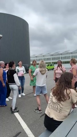 A group of women, estimated to be between eight and ten individuals, are gathered in a circle outdoors, seemingly engaged in dancing or a similar activity. The setting appears to be a paved area, possibly a parking lot, with a large, gray, cylindrical building on the left and a row of greenhouses in the background to the right. The sky is overcast. The women are dressed in casual attire, ranging from dresses to jeans and tops. Some are actively dancing, with arms raised and bodies in motion, while others are observing. The focus is on the central figures who are leading the activity. The camera is positioned at a medium distance, capturing the group from a slightly elevated angle. The overall atmosphere seems energetic and social.