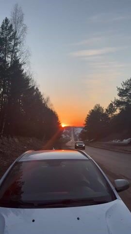 The video shows a white car parked on the side of a road, with the camera pointed towards a sunset at the end of the road. Tall trees line both sides of the road, and a few houses can be seen in the distance. There is some snow on the ground. Another car is driving down the road toward the camera.