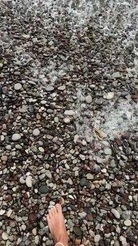 The video opens with a close-up view of a pebble beach, with the edge of the ocean visible. A person's bare foot is in the lower left corner of the frame. As the video progresses, waves begin to crash onto the shore, covering the pebbles in foam. The camera then pans out slightly to show more of the ocean and the sky, revealing a bright, sunny day. The waves continue to roll in, creating a peaceful and serene atmosphere. The video concludes with a wider shot of the beach and the ocean, with the waves receding.