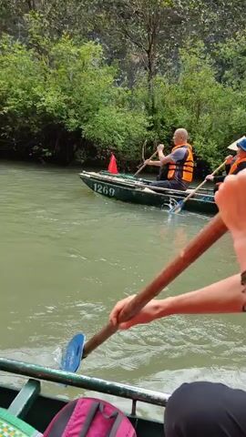 A woman is paddling a boat on a river, with lush green trees lining the banks. Two other boats are visible ahead, each carrying two people, one of whom is paddling. Everyone is wearing a life vest. The woman in the foreground turns to look at the camera. She is wearing sunglasses on her head, a patterned scarf, and bracelets on her wrist. A pink backpack can be seen in the bottom left of the frame.