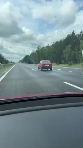 A red car is driving on a highway. The car is in the right lane and is in front of the camera car. The highway is wet, indicating it may have rained recently. There are trees on the right side of the road and a cloudy sky above.