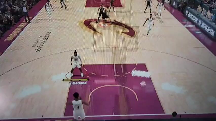 The video shows a basketball game being played on a court with the Cleveland Cavaliers logo at center court. Players wearing white jerseys are on offense against players in black jerseys. One of the white jersey players, labeled as Garland, has the ball near the top of the key. The arena is filled with spectators. The court has advertisements for Rocket Mortgage FieldHouse and Tissot.