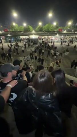 A crowd of people is walking on a staircase. Some people are walking up the stairs while others are walking down the stairs. The staircase appears to be outside at night. The environment is lit by overhead lights. Some people are holding phones.