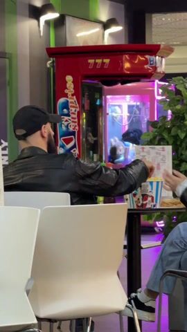 A man in a black leather jacket and black baseball cap sits at a table in what appears to be a restaurant or entertainment venue, seen from behind. He is looking at a menu in his hands and occasionally reaching into a popcorn bucket on the table. There are other food items on the table. To the right of the man, another person's legs and blue jeans are visible. Behind the man is a red claw machine with a "777" sign on top. The claw machine is lit with pink and blue lights, and inside, there are stuffed animals. A plant is positioned to the right of the claw machine, and the wall is painted with green and gray stripes.  Two lights are visible on the upper left wall above the machine.
