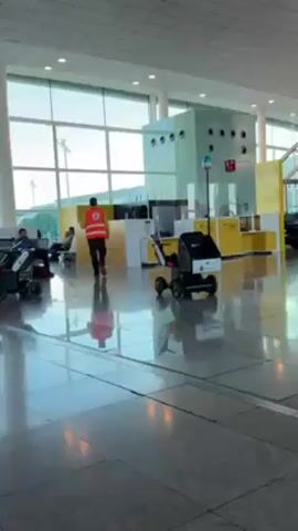 The video shows the interior of an airport terminal. The floor is shiny and reflective. A man in a bright orange vest with a wheelchair symbol on the back walks away from the camera. Several small black electric mobility vehicles with seats move across the floor. These vehicles have lights and a small screen. People are seated in the background. The terminal has large windows letting in daylight.