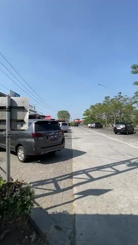 The video pans across a bright, sunny outdoor scene with a clear blue sky. The foreground features a grey SUV parked on a paved area with a brick-patterned border. To the left, a metal signpost is visible, casting a shadow. Beyond the SUV, a road stretches into the distance, lined with trees on the right side. Several other vehicles are present on the road, including a white minivan, a silver car, and two darker colored cars further down. Red structures, possibly part of a building or stalls, are visible on the left side of the road. The overall impression is that of a rest stop or a service area along a highway.