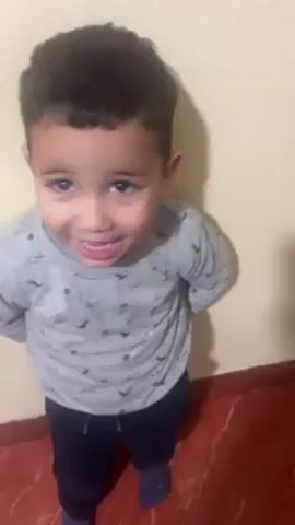 A young boy with dark, curly hair stands against a cream-colored wall. He is wearing a grey long-sleeved shirt with a pattern of small black dinosaur silhouettes and dark-colored pants. The boy smiles at the camera, occasionally glancing to his right. The floor is covered in red tiles.