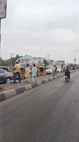 The video shows a street scene in what appears to be an urban area. On the right side of the road, several yellow auto rickshaws, also known as tuk-tuks, are parked along the curb. People are walking on the sidewalk next to them, and some are interacting with the rickshaws. Cars and motorcycles are moving along the road. A building with a billboard advertising "AA RANO LUBES LTD" is visible in the background. The sky is overcast.
