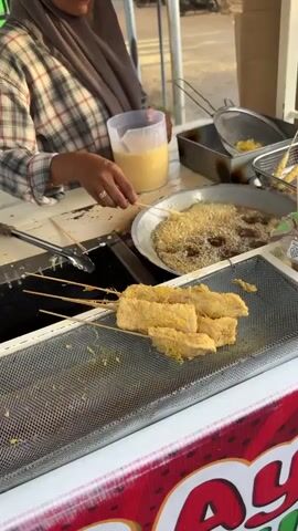 A woman wearing a hijab and a plaid shirt is standing behind a food cart. She is holding a skewer with food on it and dipping it into a pan of hot oil. In front of her, there is a metal rack with several skewers of fried food. The food appears to be tofu coated in batter. There are also metal tongs and a container of batter on the cart. The background is blurry, but it appears to be an outdoor setting with a street and some vehicles. The lighting suggests it is daytime.