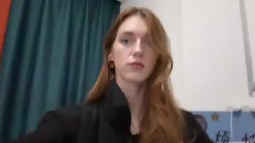 A woman with long, reddish-brown hair stands in front of a camera. She is wearing a black jacket and small earrings. Behind her is a teal curtain and a white wall with electrical outlets. She touches her hair and then smiles slightly.