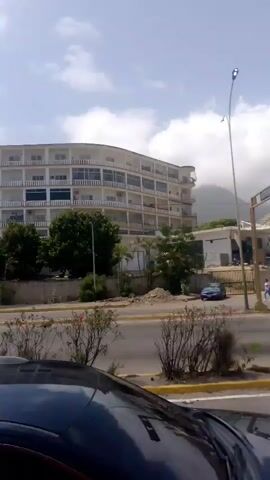 The video captures a street scene in Caraballeda, Venezuela. A multi-story building with a curved facade, likely a hotel or apartment complex, dominates the background. The building is light-colored and features many balconies and windows, suggesting it's a warm climate. Adjacent to it, several smaller structures and a billboard are visible. A body of water, possibly the sea, can be inferred from the mountainous terrain and the coastal location of Caraballeda.

In the foreground, a street with multiple lanes is depicted. Cars are driving on the road, including a blue sedan and a white truck. The truck has a distinctive logo on its side, reading "HIELO HIELO" in a circular design with blue lettering. This suggests the truck is involved in ice delivery. A traffic light and a lamppost are also present.

The weather appears to be overcast with some clouds, but the overall lighting suggests it is daytime. The presence of greenery, including trees and bushes, indicates a well-vegetated area. The scene seems to be one of a typical day in a coastal town, with ongoing traffic and everyday activities.
