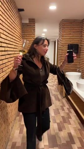 A woman stands in a bathroom, holding a glass of white wine in one hand and a phone and small purse in the other. She wears a brown jacket with flared sleeves and a tie belt, a brown top, and black pants. The bathroom has brick walls and a light-colored tiled floor. There are white sinks along one wall. The woman walks toward the camera, exclaiming "Beautiful bathroom, you know?". She gets very close to the camera lens, causing the image to blur.