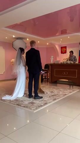 Two individuals, a bride in a white wedding dress with a veil and a groom in a dark suit, stand facing a registrar at a wooden desk. The registrar, a woman with dark hair and a striped jacket, speaks into a microphone. Behind her, a flag and a coat of arms are visible on the wall. The room is decorated with pink walls and a pink reflective ceiling. A rug with a patterned design is on the floor. The scene appears to be a civil wedding ceremony.