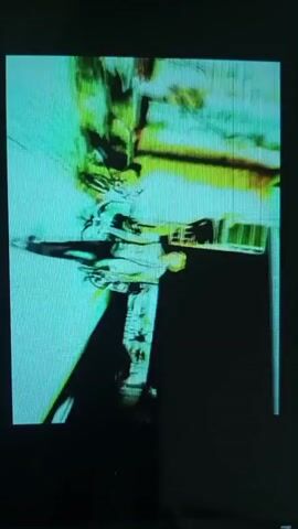 The video displays a distorted, glitchy image of what appears to be a person's face. The image is highly pixelated and fragmented, with bright green and yellow colors dominating the frame. The person's features are difficult to discern due to the distortion. The background is dark and indistinct. The overall impression is one of digital degradation or manipulation.