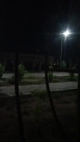 This is a dark, shaky video clip of an outdoor scene at night. A bright streetlight is visible in the upper right corner, casting light on a path and some small bushes. There are dark, vertical posts in the foreground, and a low-lying fence or barrier is also visible. In the background, a building with lights on can be seen, along with distant streetlights and the headlights of a vehicle. The overall impression is of a deserted, dimly lit area, possibly a park or campus.