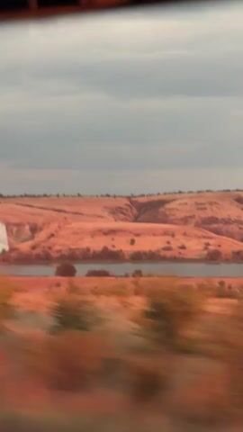 This media file depicts a blurry, fast-moving scene through a window, likely from a vehicle. The primary subject is a vast, rolling landscape bathed in the warm, orange-pink hues of what appears to be sunset or sunrise. The terrain is characterized by dry, reddish-brown hills or bluffs, with evidence of arid vegetation and sparse trees. A body of water, possibly a river or lake, is visible in the mid-ground, reflecting the warm light. The sky is overcast with gray clouds, suggesting a possibly cool or transitional weather. The movement of the camera creates a sense of journey and transience, blurring the details of the landscape into streaks of color. There are no visible people or specific objects that stand out, only the encompassing natural environment. The overall mood is somewhat impressionistic and atmospheric, emphasizing the vastness and muted beauty of the rural setting. The location context suggests this scene is in Russia, specifically near Ferma Sovkhoz Balykleyskiy. The time of day is indicated by the lighting to be early morning or late evening, and the weather appears to be cloudy.