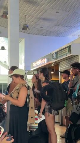 Several people are standing in line inside a building. The woman in the middle wears a leopard print outfit and a black backpack. A man with a black baseball cap is behind her. To the left, a woman in a black dress and light green hat looks at her phone. Overhead, there are hanging light fixtures. A coffee shop is visible behind the people. The scene is bright.