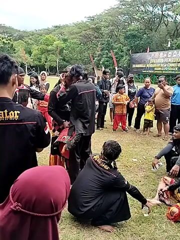 The video shows an outdoor event on a grassy field, where a group of people are gathered around what appears to be a traditional performance. Several individuals wear black shirts with the word "Predator" emblazoned on the back in gold lettering. In the center of the group, a person in a black outfit appears to be assisting someone on the ground, who is lying down and possibly wearing a traditional costume. There is a backdrop of trees and a banner in the distance. Some of the spectators are holding phones, possibly recording the event. The overall atmosphere seems to be one of cultural celebration or performance.