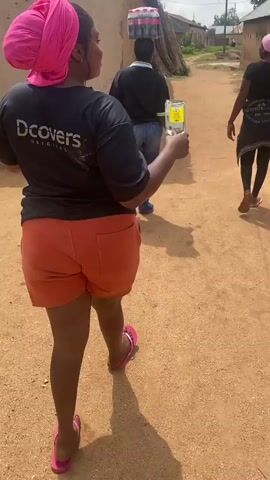 The video shows the backs of two women walking on a dirt path. The woman in the foreground is wearing an orange pair of shorts and a black t-shirt with the logo "DCovers Original" on the back. She has a pink headscarf and pink flip-flops. In her right hand, she is holding a clear plastic cup with a yellow liquid and a straw. Behind her, a second woman is walking away from the camera. She is wearing a black t-shirt and a pink headscarf. Another person is visible in the distance, also walking away. The path appears to be in a rural or semi-rural setting, with simple buildings visible in the background. The lighting suggests it is daytime.
