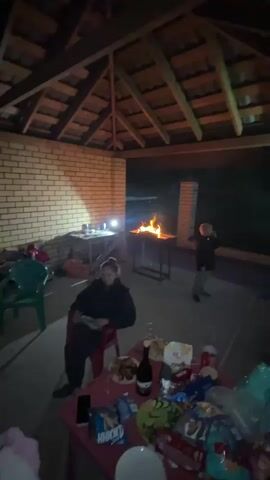 A group of people are gathered in an outdoor covered patio area at night. A fire is burning in a grill in the background. There is a table in the foreground with food and drinks on it. Two women are sitting on chairs, and a man is standing. The scene is lit by the fire and some artificial lighting.